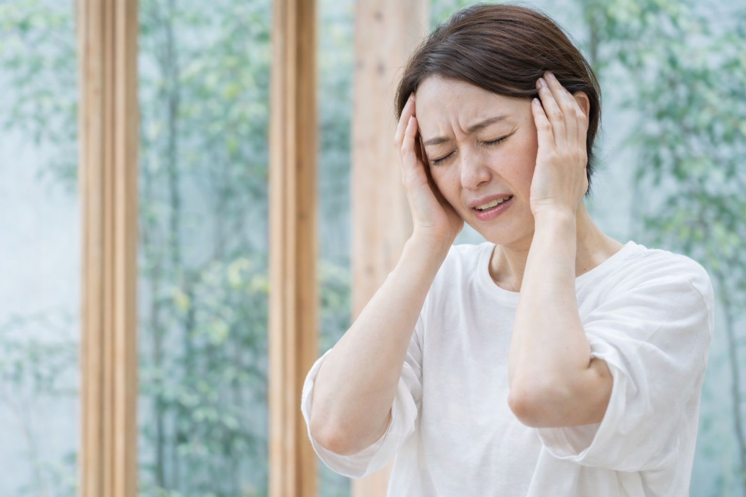 頭を両手で押さえ、つらそうな表情を浮かべる女性。頭痛やめまい、肩こりからくる不調を訴えている様子