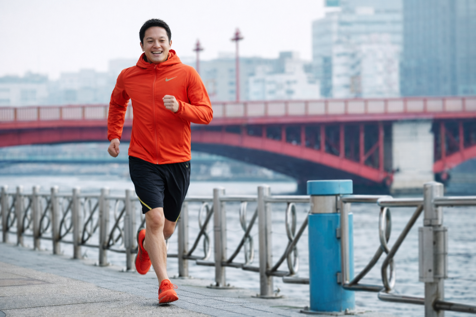 「川沿いの遊歩道を笑顔でランニングする男性。オレンジのウェアを着て軽快に走っている様子」
