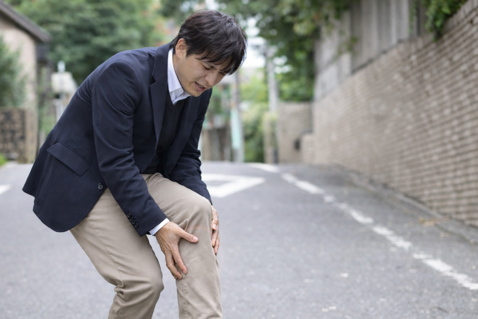 「ふくらはぎに手を当てて痛みを感じているスーツ姿の男性。坂道で足の不調に苦しんでいる様子。」