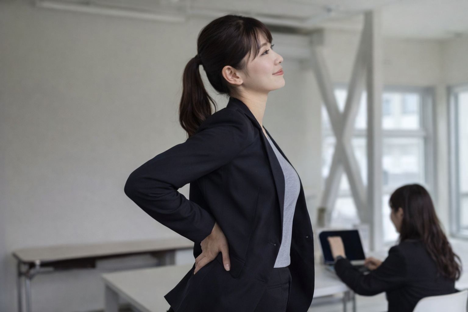 「腰に手を当てて反り腰の違和感を感じているビジネスウーマン。デスクワーク中の姿勢不良が原因の腰の張り」