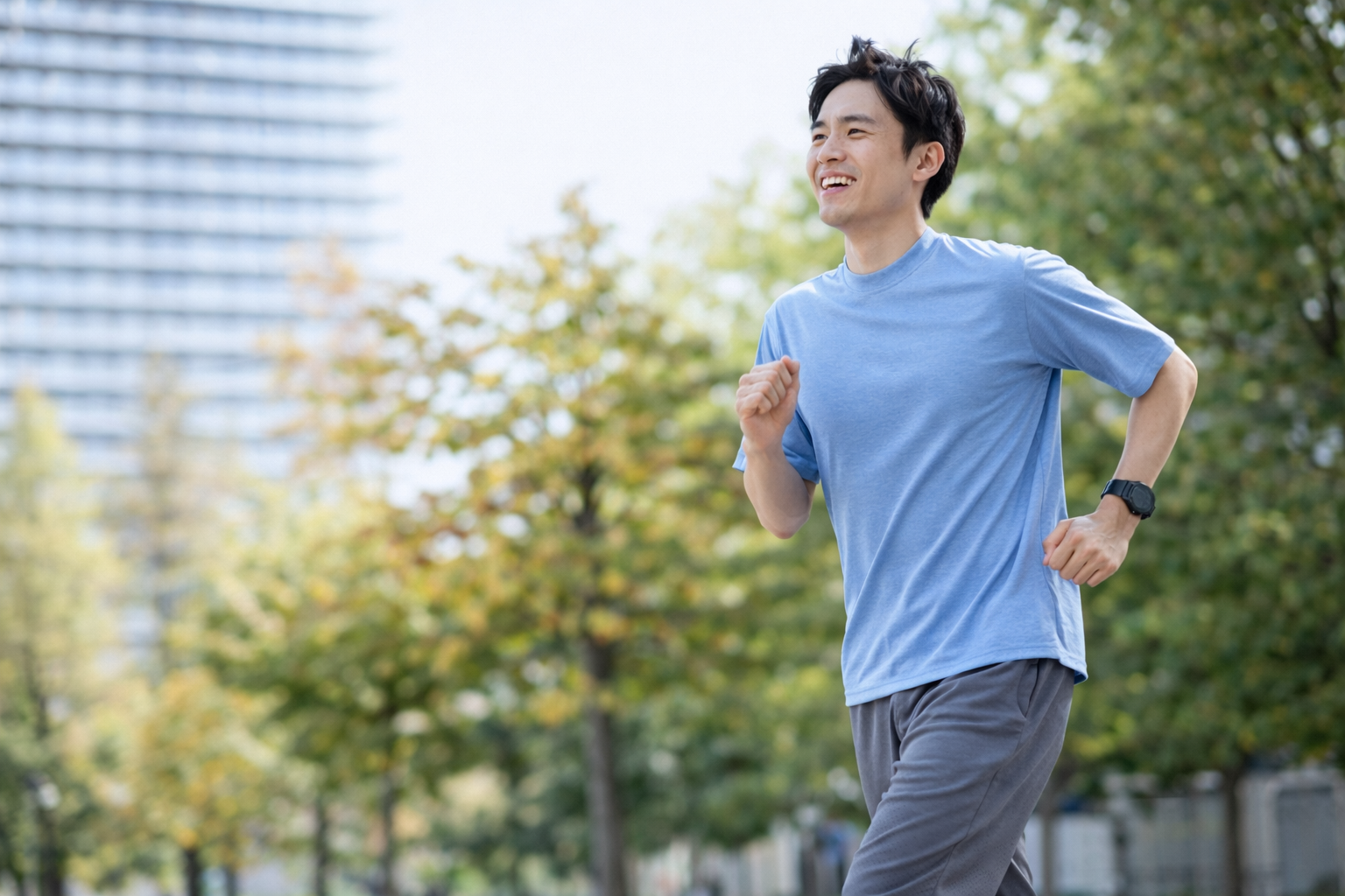 「公園の遊歩道を笑顔でランニングする男性。青いTシャツとグレーのパンツを着用し、快調に走っている様子。」
