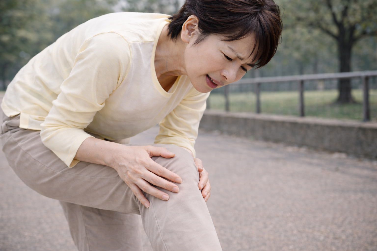 「屋外で膝を押さえながら痛みを訴える中年女性。膝関節の不調や加齢による膝痛のイメージ。」