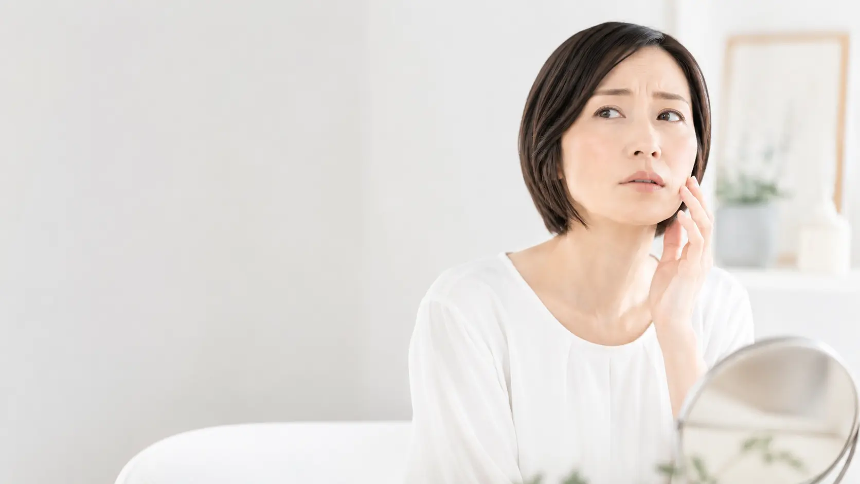 鏡を見ながら顔のたるみやフェイスラインを気にする40代女性。美容鍼による肌悩みや表情の変化をイメージした画像。