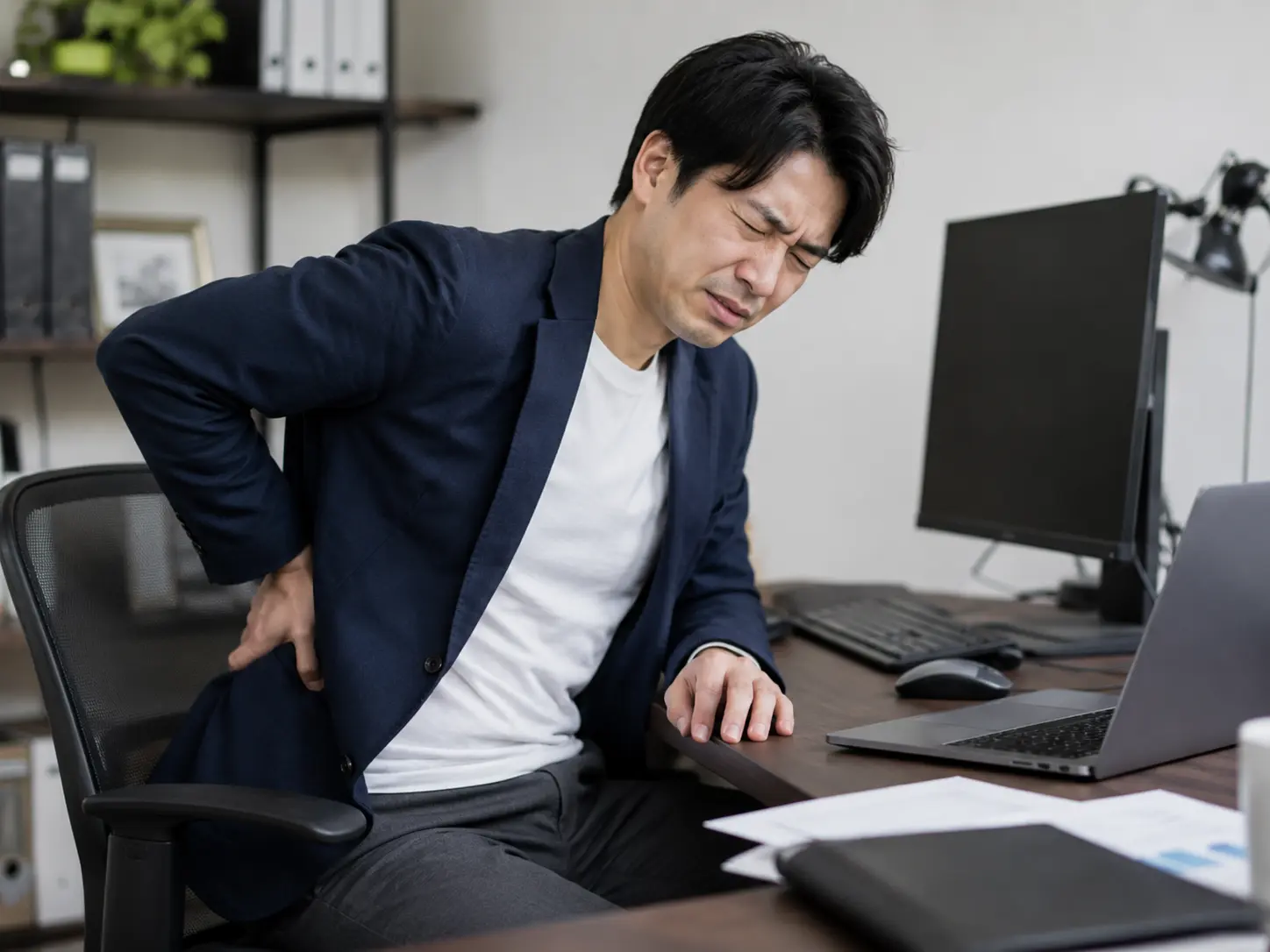 デスクワーク中に椅子に座ったまま腰を押さえ、痛みを感じている日本人男性。日野市で整骨院を探している腰痛の方へ向けたブログ用画像