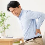 デスクワーク中に椅子へ座ったまま腰を押さえ、痛みを感じている日本人男性。日野市で整骨院を探している腰痛の方へ向けたブログ用画像