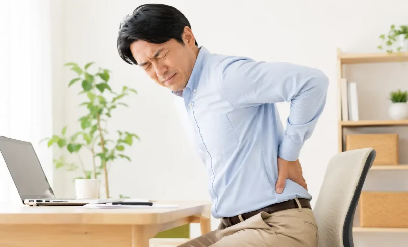 デスクワーク中に椅子へ座ったまま腰を押さえ、痛みを感じている日本人男性。日野市で整骨院を探している腰痛の方へ向けたブログ用画像