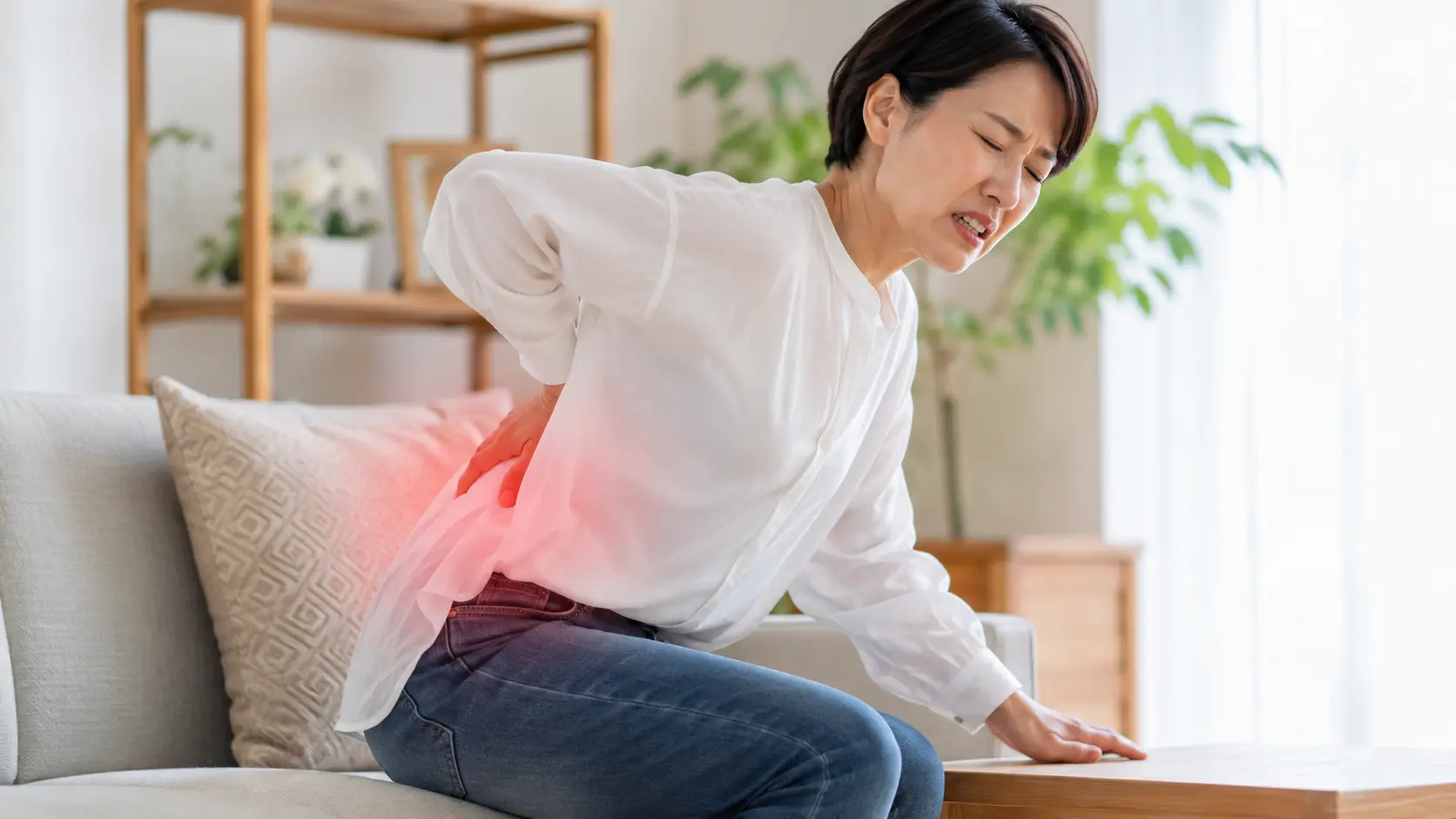 ソファから立ち上がろうとして腰に手を当て、強い腰痛を感じている女性
