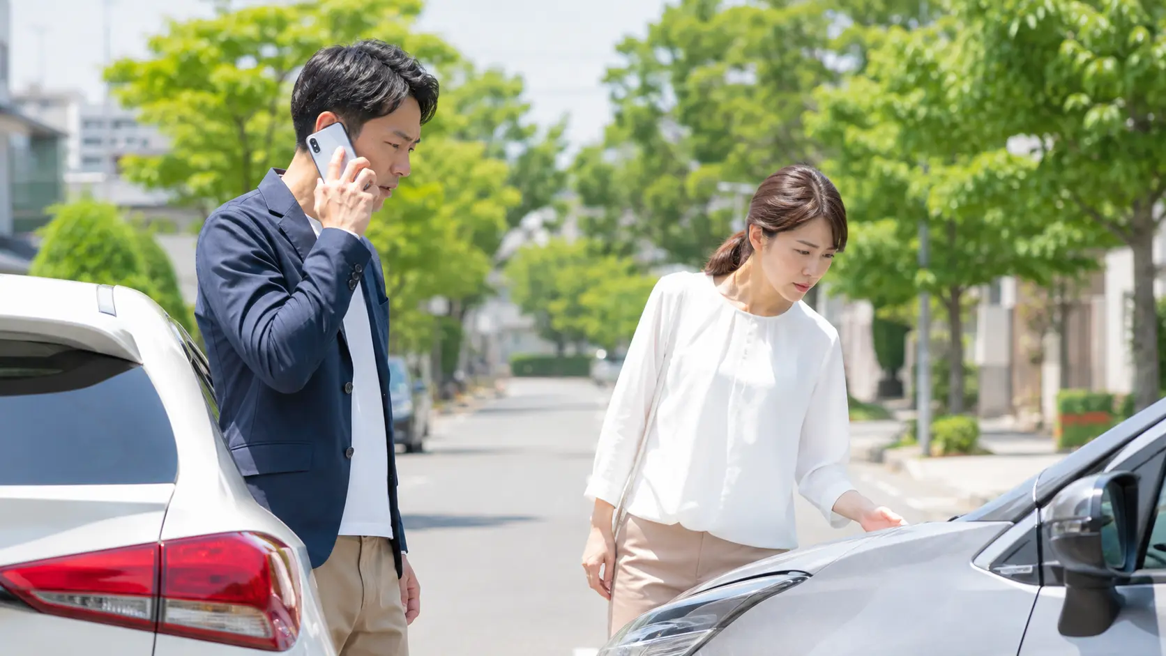 交通事故直後に車の状態を確認しながら、スマートフォンで警察や保険会社へ連絡する男女