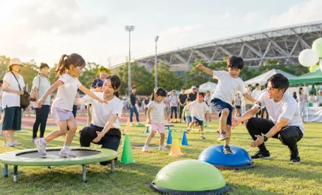 八王子富士森競技場の屋外イベント会場で、子どもたちがトランポリンやバランスボールを使った運動体験を楽しみ、からだケア整骨院のスタッフがサポートしている様子を表したアイキャッチ画像。家族連れや来場者が周囲で見守り、地域イベントのにぎやかで明るい雰囲気が伝わる。