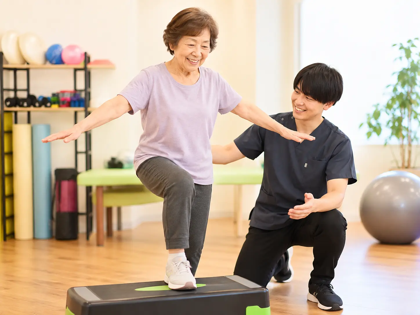 スタッフのサポートを受けながら、台を使って片脚立ちのバランストレーニングを行うシニア女性の様子。